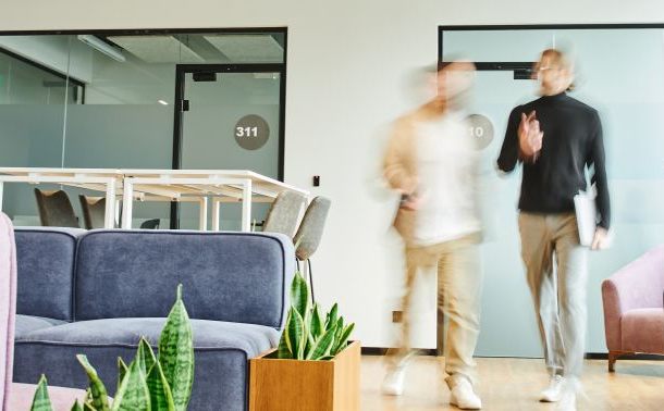 Two people walking past seating areas in an office space.